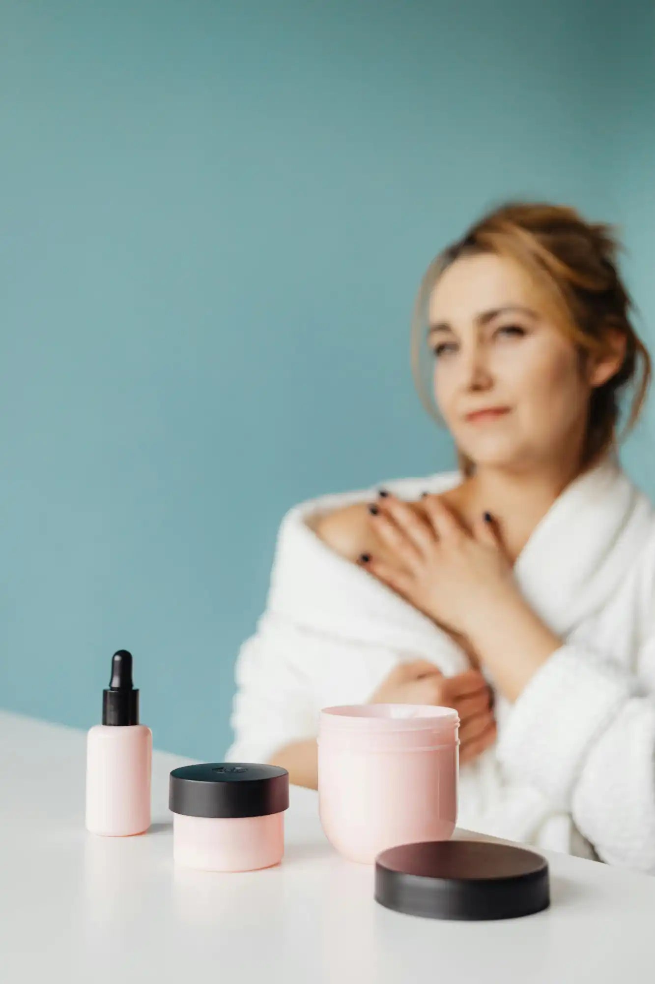 Une femme en peignoir blanc avec divers produits de soins de la peau sur une table.