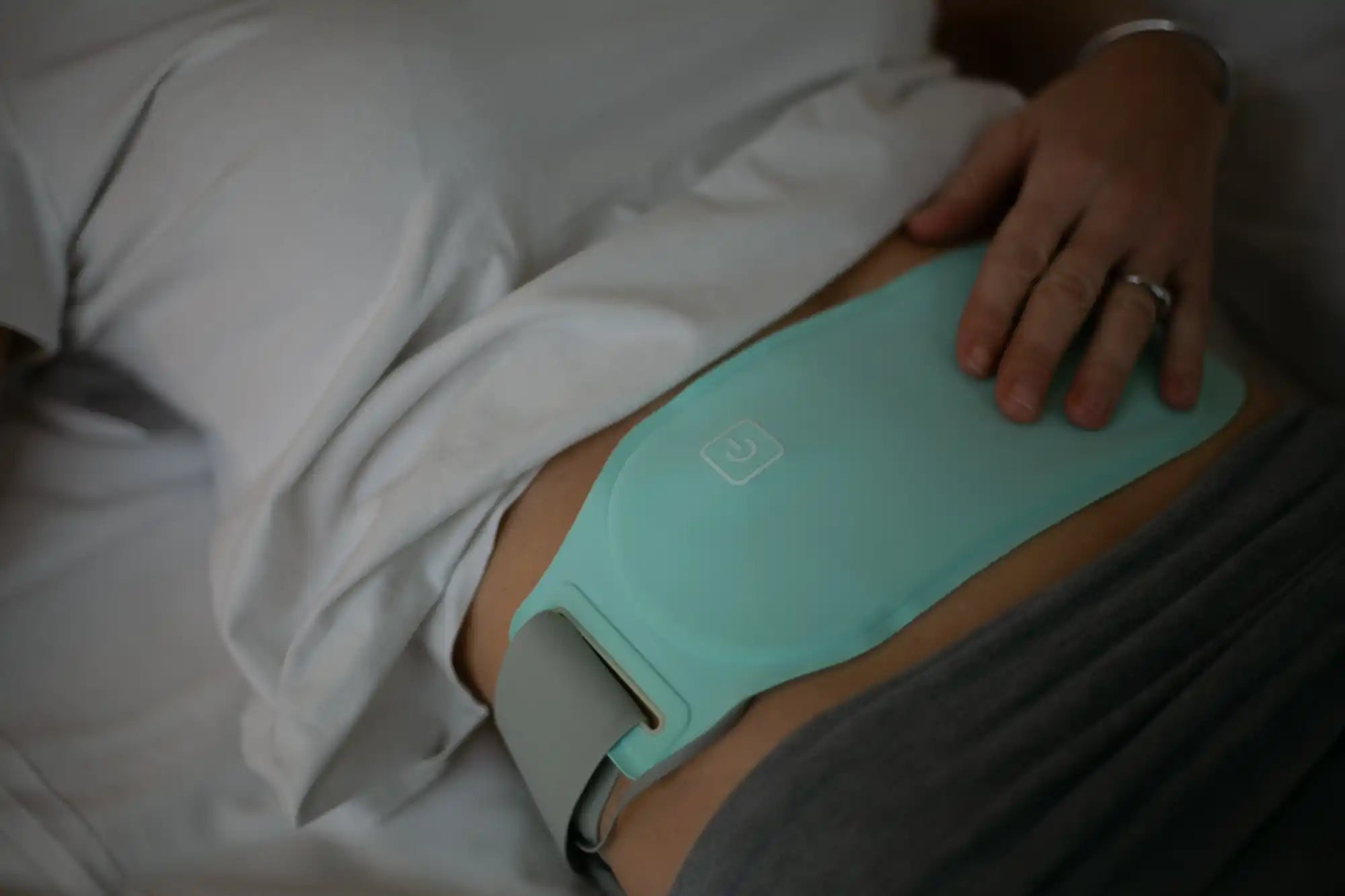 Une bouillotte souple de couleur bleu-vert clair avec une bandoulière grise réglable, conçue pour soulager l'abdomen ou le dos.